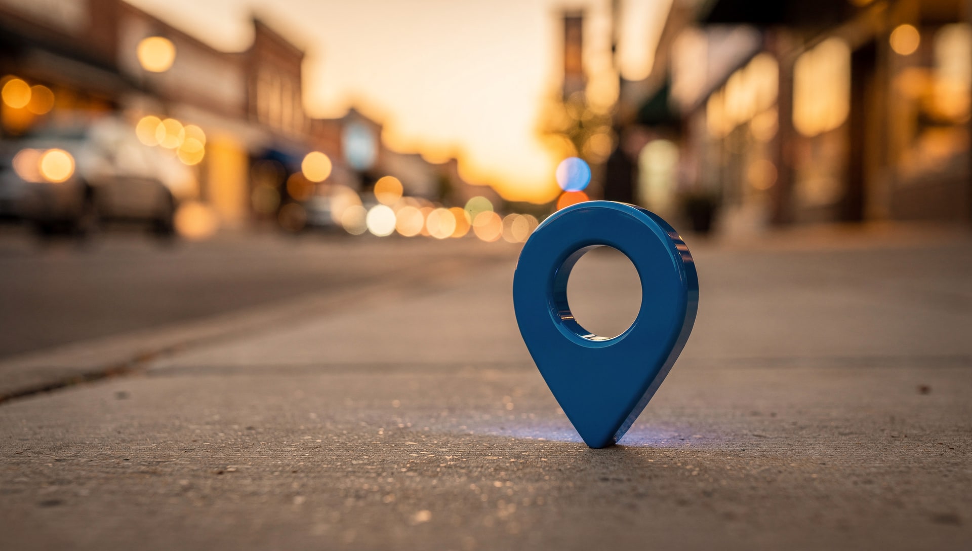 Blue map pin standing on the sidewalk of a small-town main street with shop lights glowing behind it